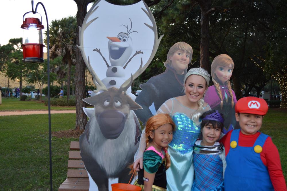 Young woman dressed as Elsa poses for picture with girl dressed as Merida from Brave, girl in blue dress and boy dressed as Mario in front of Frozen character cutouts